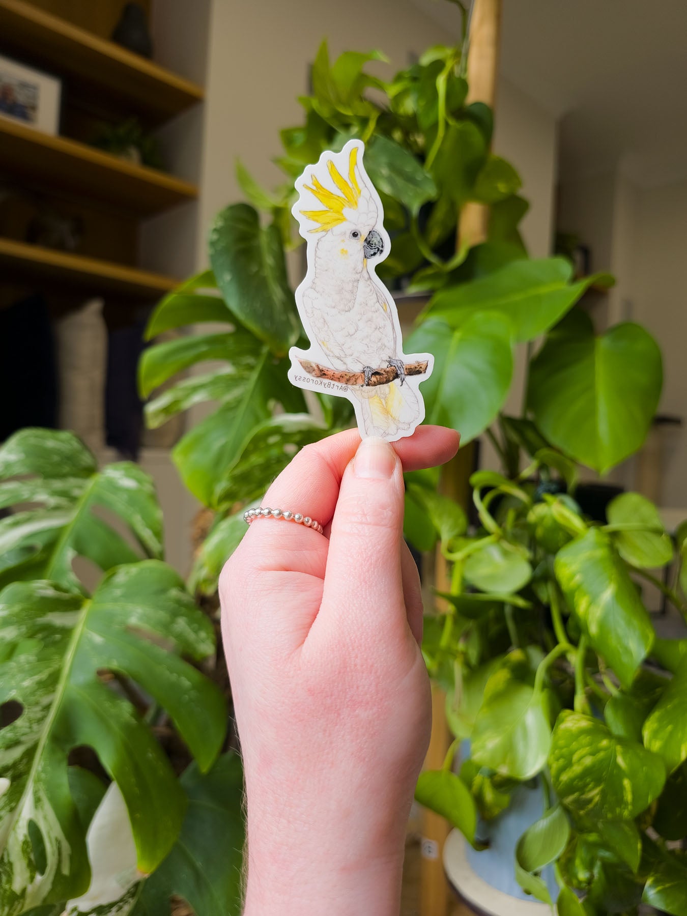 Hand holding a temporary tattoo of a white parrot with a yellow crest against a background of green plants.